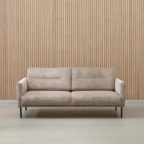 Beige tygsoffa med svarta ben framför en vägg med vertikala träslister.