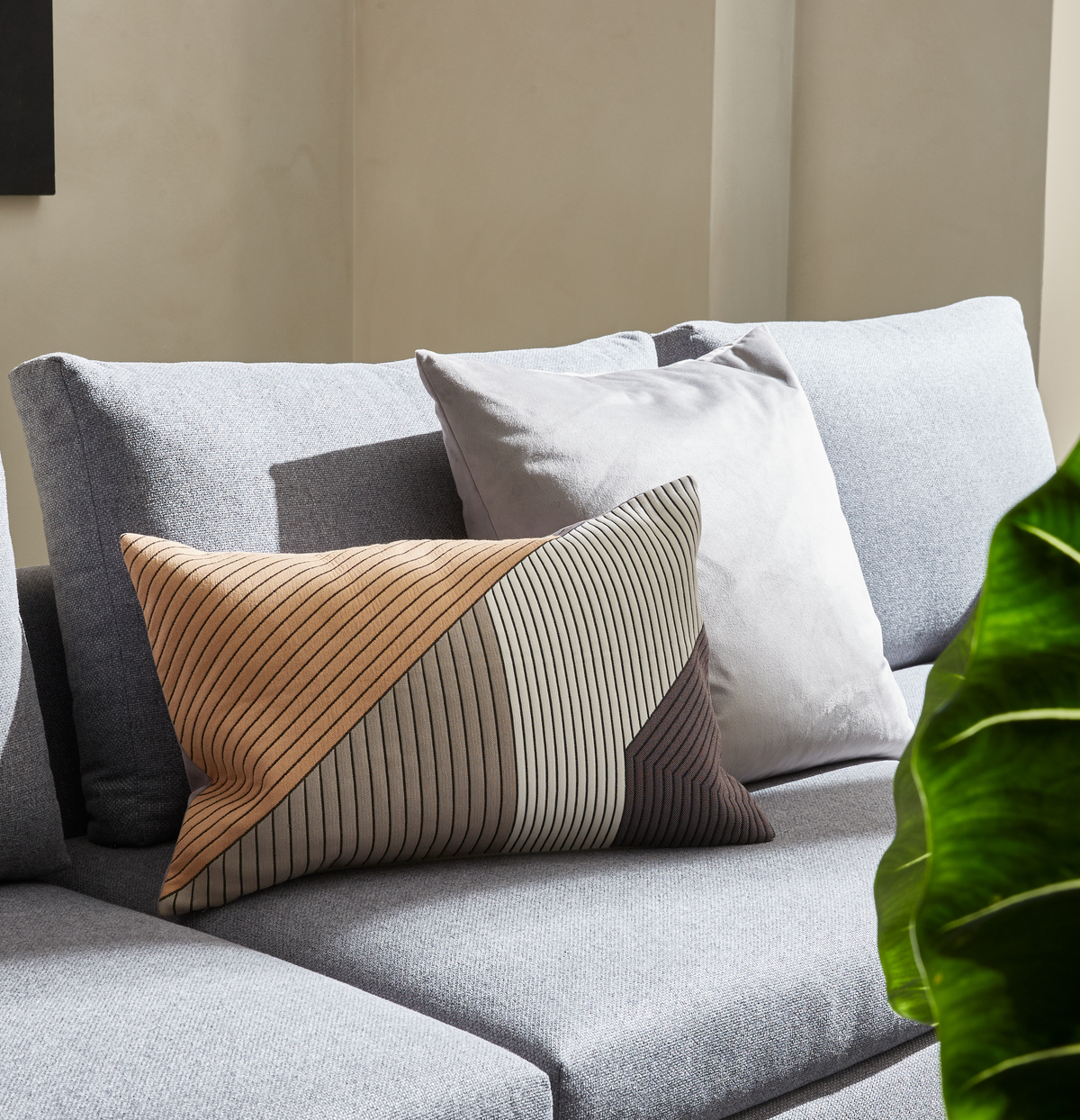 Ljusgrå soffa med två kuddar – en ljusgrå och en rektangulär med geometriskt mönster i beige, grått och brunt – i solbelyst vardagsrum med grön växt i förgrunden.