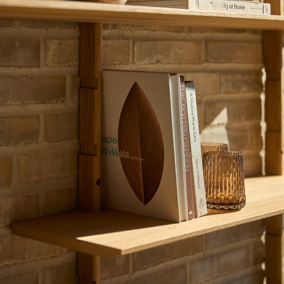 Trähylla mot tegelvägg med böckerna ”Tapio Wirkkala”, ”The Potted Garden”, ”A Modern Way to Live” av Matt Gibberd och ”Simplicity at Home”, bredvid ett bärnstensfärgat glas i varmt dagsljus.