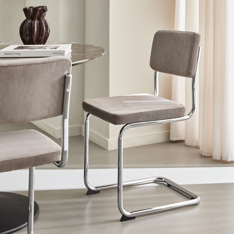 Modern karmfri stol i beige manchester med kromad fribärande ram, intill runt marmorbord med en mörk vas och böckerna ”barefoot living” och ”HOTEL Weekend”.