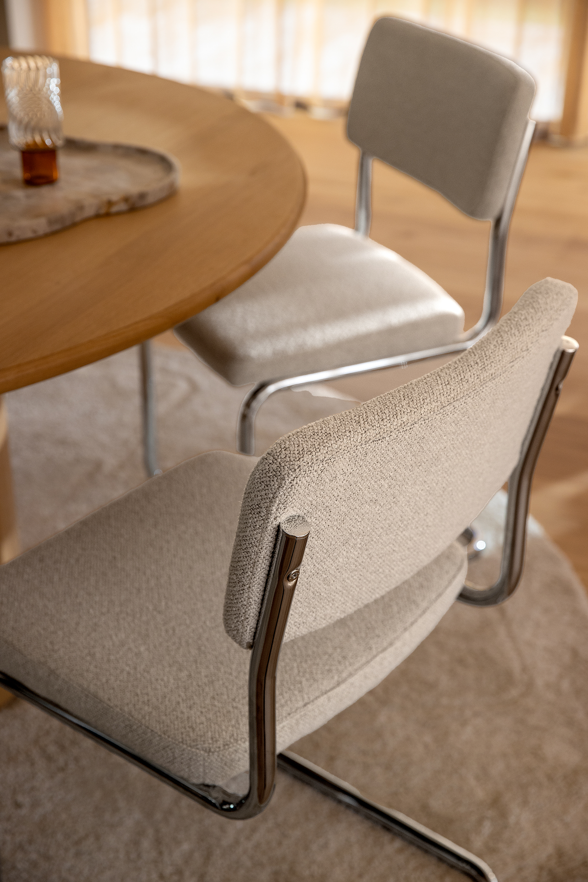 Närbild på två beige stoppade cantilever-stolar med kromställ vid ett runt matbord i trä, i mjukt dagsljus på en neutral matta.