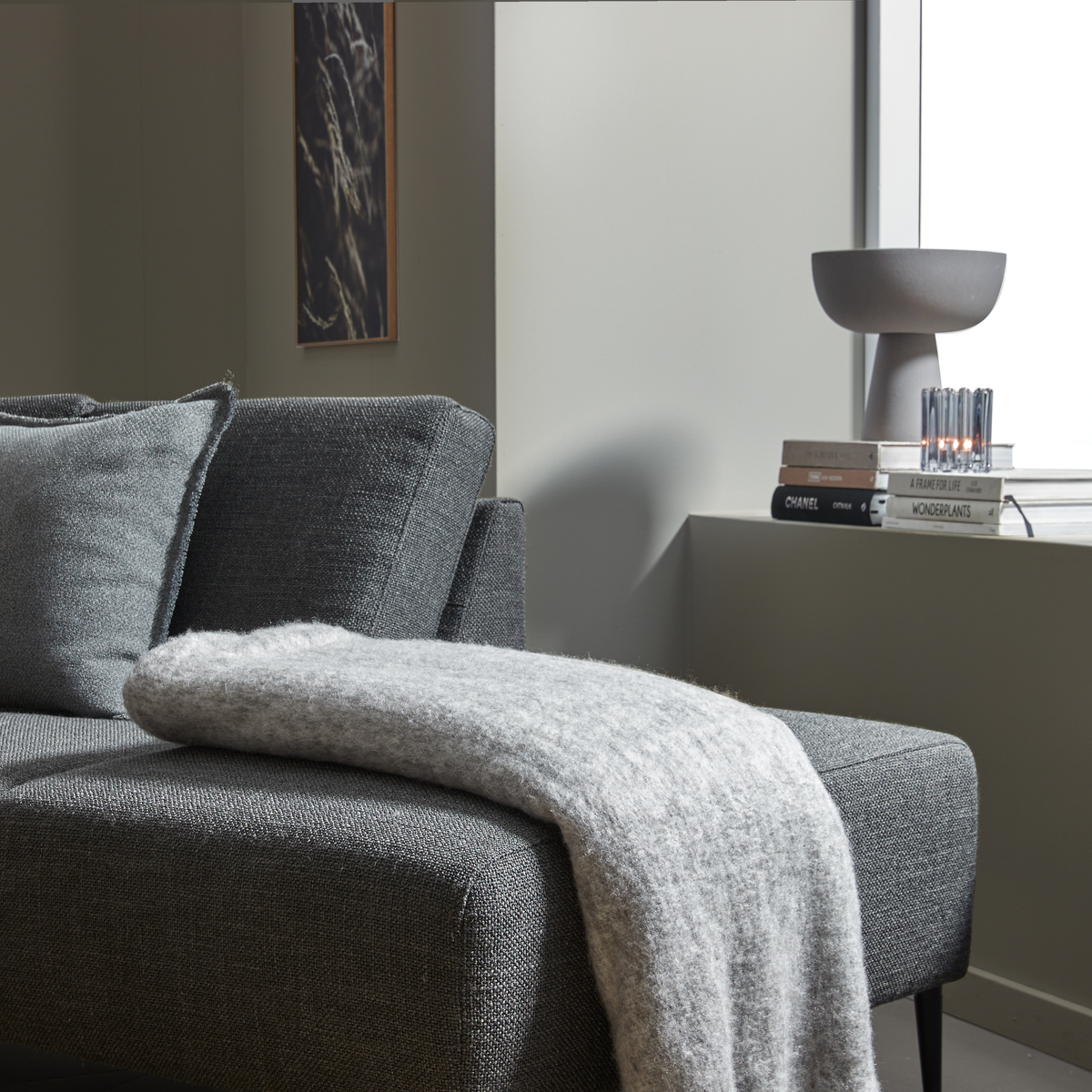 Närbild av en grå soffa med ljusgrå pläd; på fönsterbrädan syns en grå bordslampa, ett litet ljus och en bokhög med titlarna CHANEL, A FRAME FOR LIFE och WONDERPLANTS.