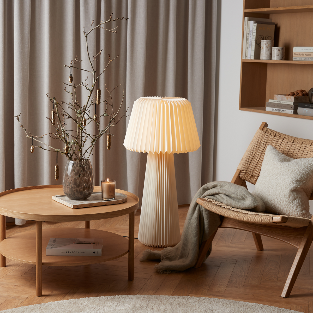 Vardagsrumshörna i skandinavisk stil med vävd loungefåtölj, kudde och filt, plisserad golvlampa och runt soffbord med vas med kvistar och ett tänt doftljus; böckerna KINFOLK HOME och New Nordic Houses syns på hyllan.