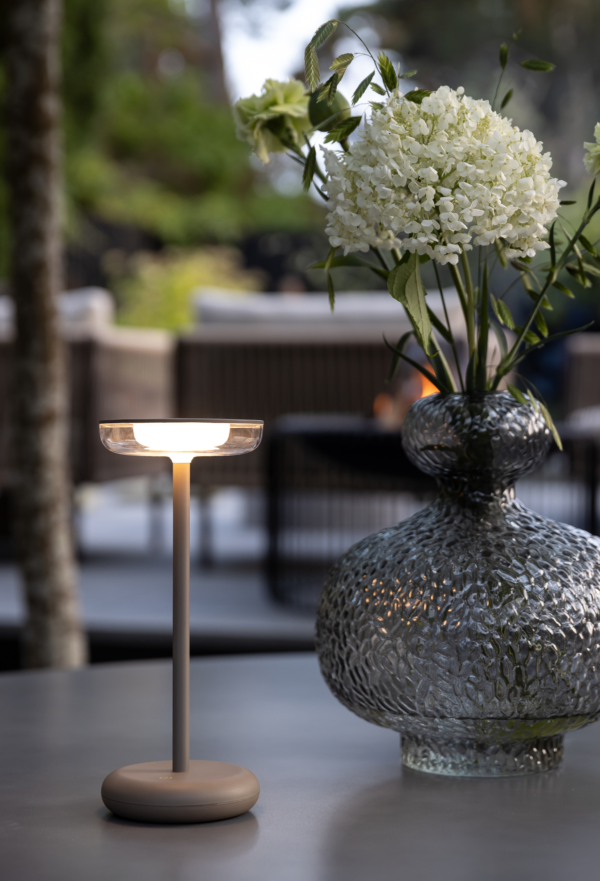 Bärbar LED-bordslampa tänd bredvid en texturerad glasvas med vita blommor på ett utebord.