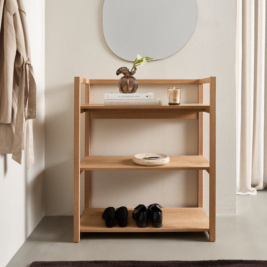 Ljus hallscen med en hylla i ek under en organisk spegel; översta hyllan har en glasvas med en blomma, en ljuslykta och böckerna The Kinfolk Home och Soft Minimal, nederst står svarta skor.