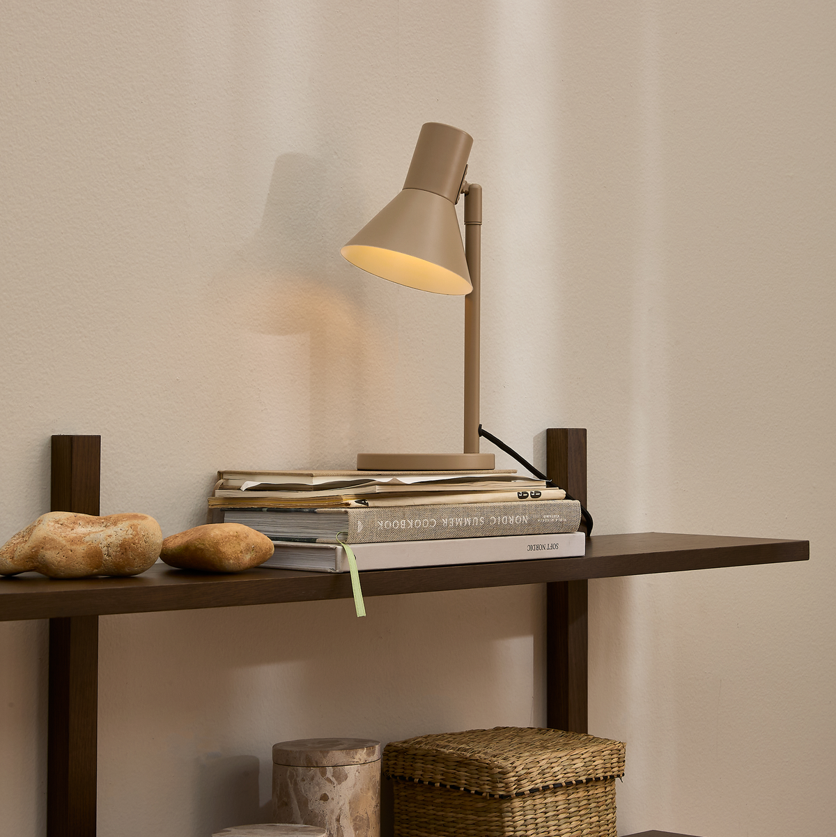Beige bordslampa på mörk trähylla med böckerna Nordic Summer Cookbook och Sky Design, naturstenar och flätad korg i lugn, minimalistisk inredning.
