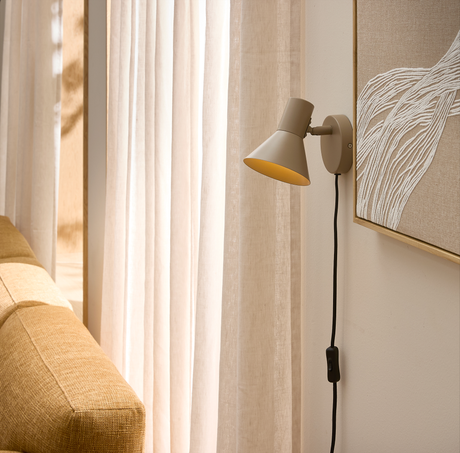 Beige vägglampa med svart sladd och brytare ovanför en gulbrun soffa bredvid ljusa gardiner i vardagsrum med mjukt dagsljus.