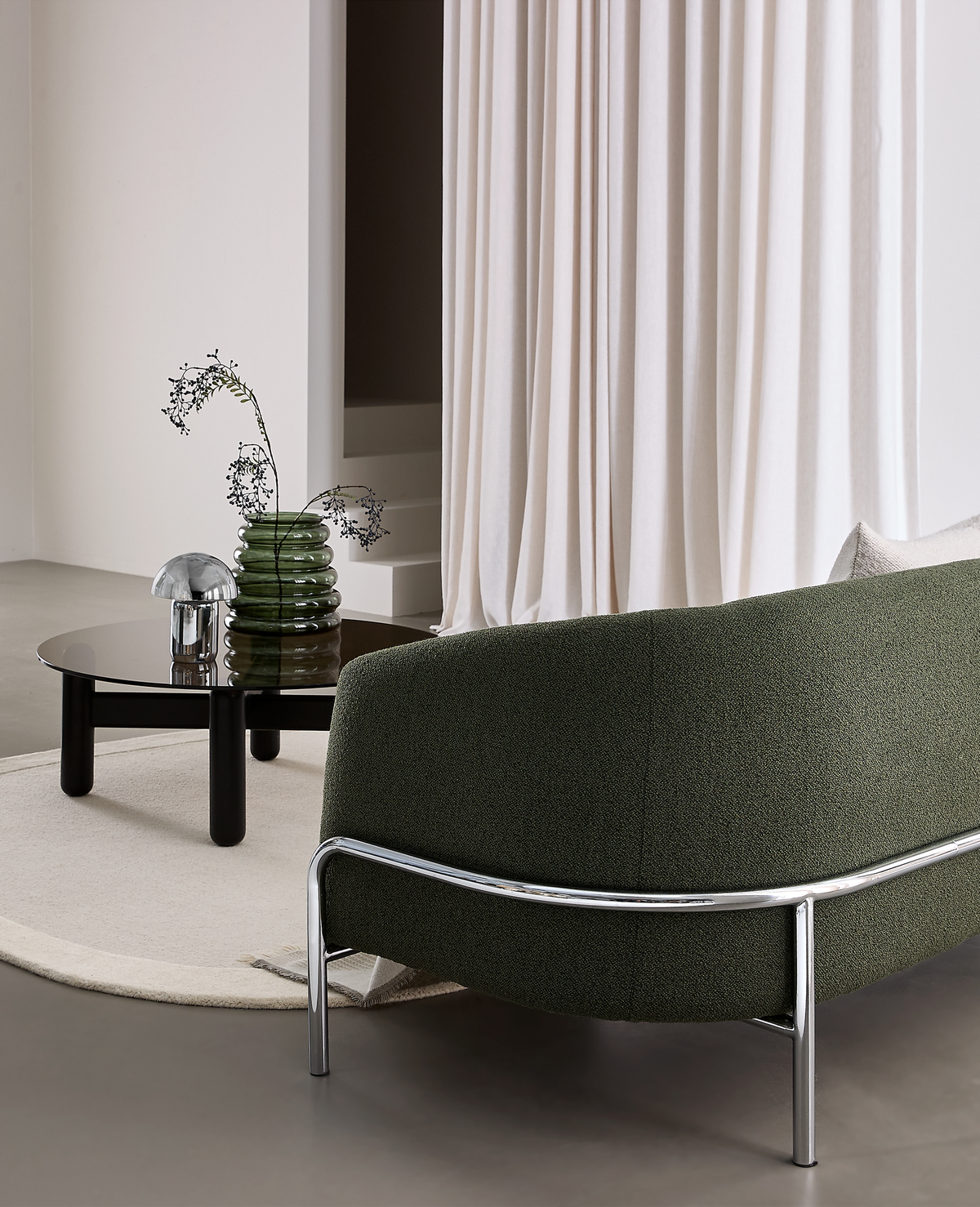 Modernt vardagsrum med mörkgrön rundad soffa med kromben, runt svart soffbord med rökfärgat glas, grön räfflad vas med kvistar och liten krombordslampa på ljus rund matta framför ljusa gardiner.