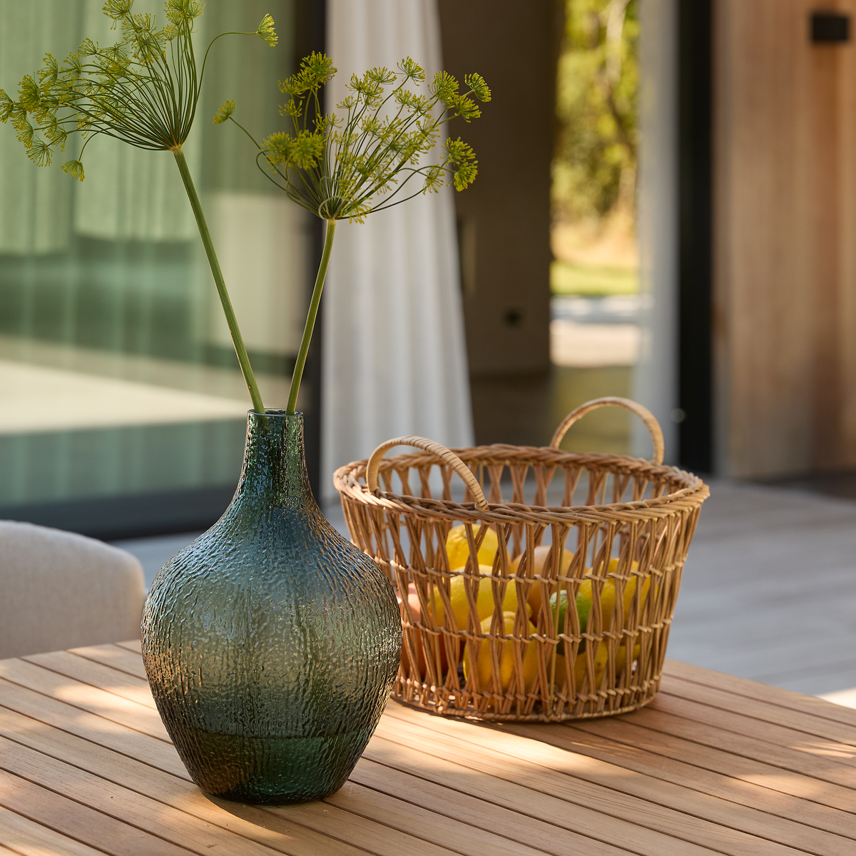 Grön texturerad glasvas med gröna stjälkar bredvid en flätad korg med citroner på ett träbord i ett soligt utrymme utomhus.