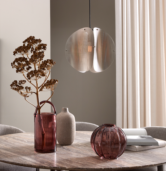 Runt matbord i trä med två vinröda glasvaser, en beige keramikvas och en torkad kvist; ovanför hänger en metallisk pendellampa, ljusa gardiner i bakgrunden.