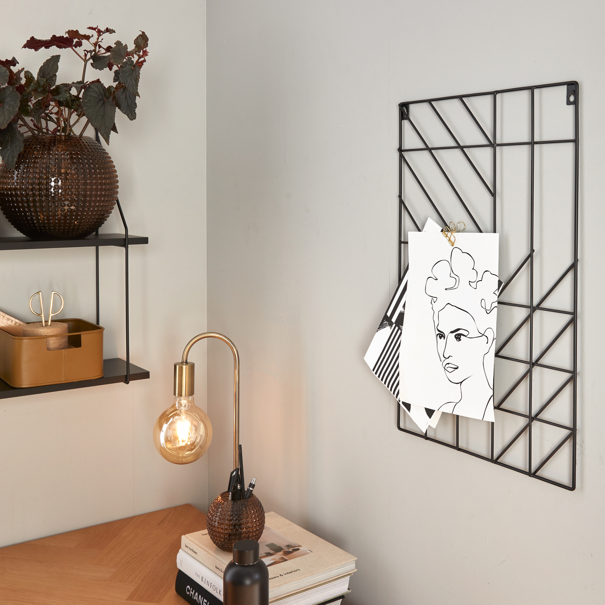 Hemmabyråhörna med träskrivbord, svart metallgaller med uppklämda illustrationer, tänd bordslampa i mässing och glas, växt i brun vas på hylla och böcker på bordet med ryggarna ”Chanel” och ”Kinfolk”.