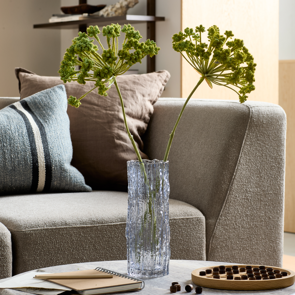 Texturerad glasvas med två gröna blomstjälkar på ett soffbord framför en grå soffa, kuddar, anteckningsbok och ett träspel i ett vardagsrum.