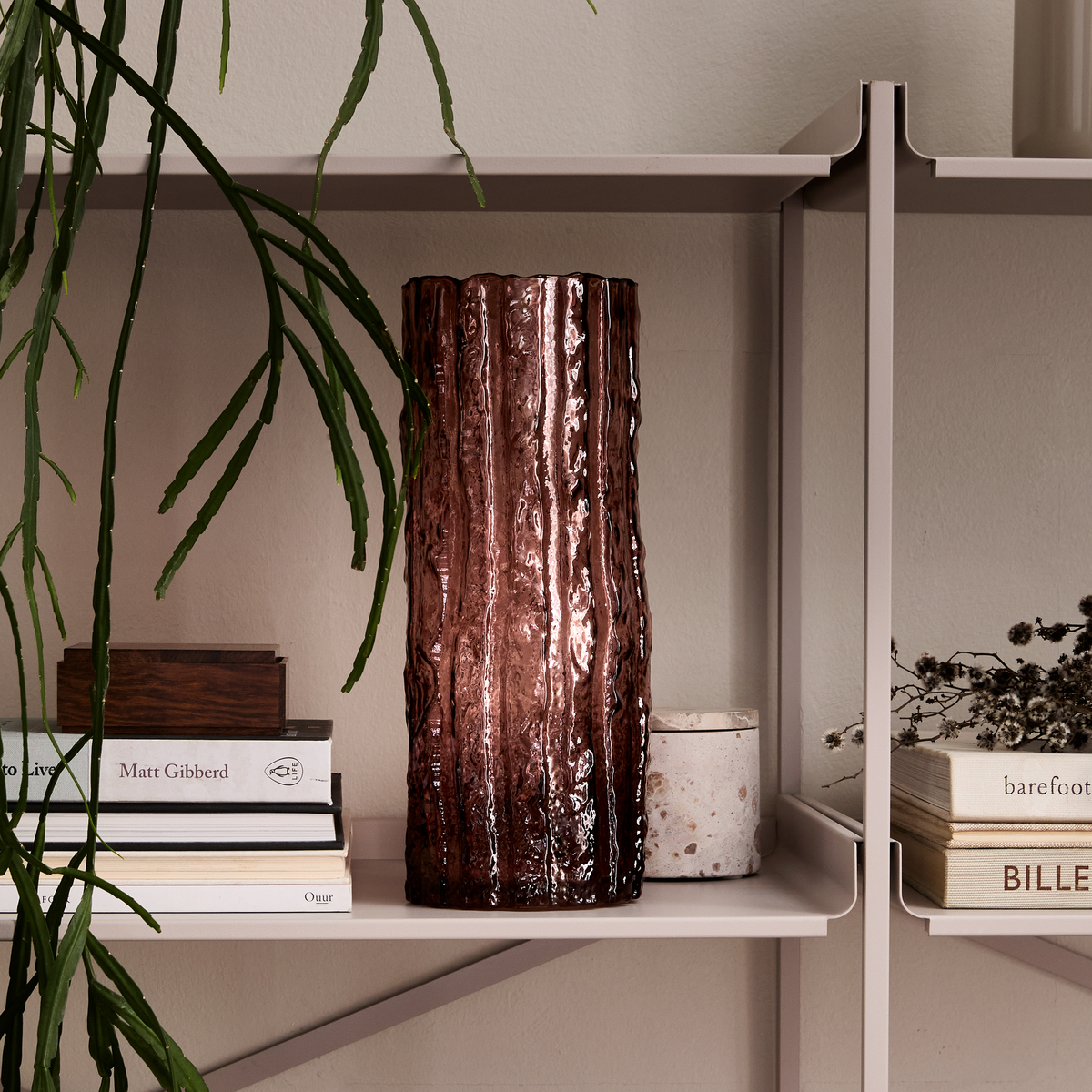 Brun, räfflad glasvas på hylla med böcker (Matt Gibberd, Ouur, barefoot) och en grön hängväxt i en lugn skandinavisk interiör.