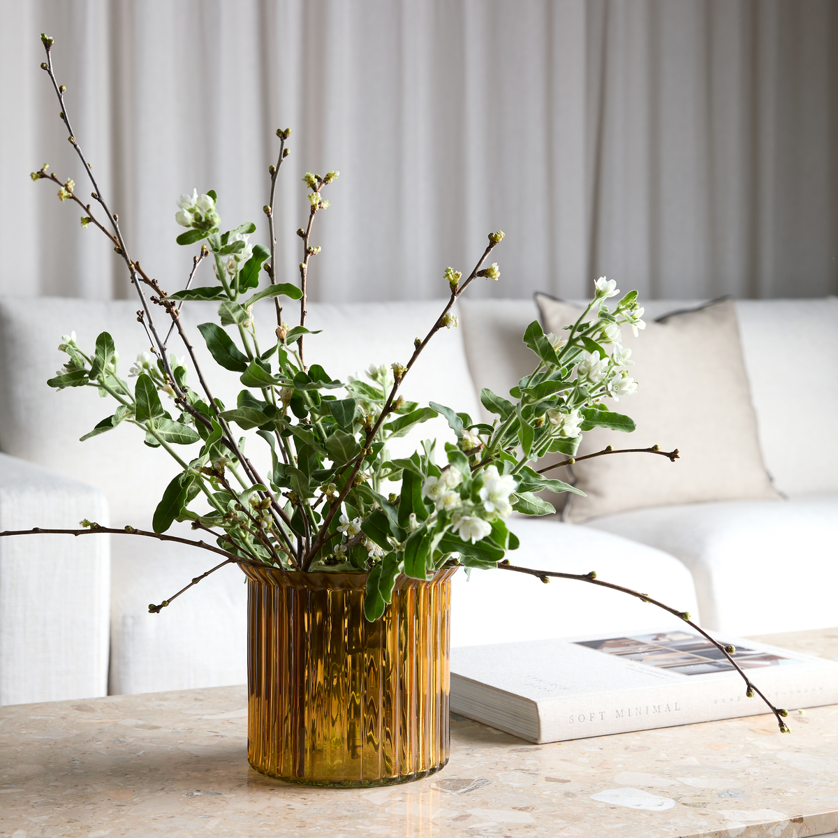 Bärnstensfärgad glasvas med gröna kvistar och vita blommor på stenbord i vardagsrum; beige kudde och boken Soft Minimal i bakgrunden.