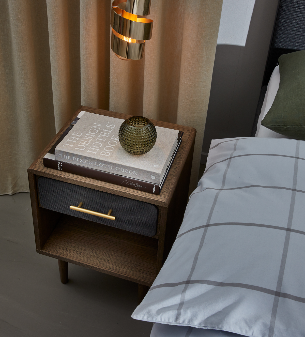 Nattduksbord i mörkt trä med mässingshandtag bredvid sängen; ovanpå ligger designböcker med titeln THE DESIGN HOTELS BOOK och en liten räfflad glasvas, med mässingspendel och beige gardiner i bakgrunden.