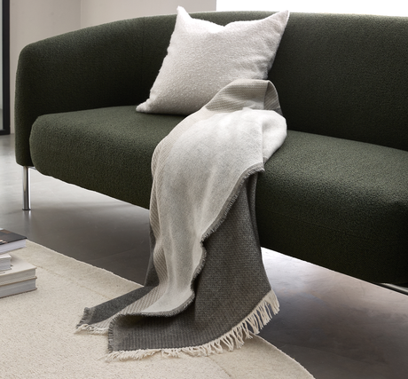Mörkgrön soffa med ljus kudde och tvåfärgad grå pläd med fransar draperad över sitsen i ett minimalistiskt vardagsrum.