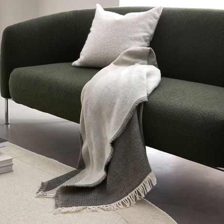 Mörkgrön soffa med vit kudde och gråvit pläd med fransar draperad över kanten, på en ljus matta i ett ljust vardagsrum.