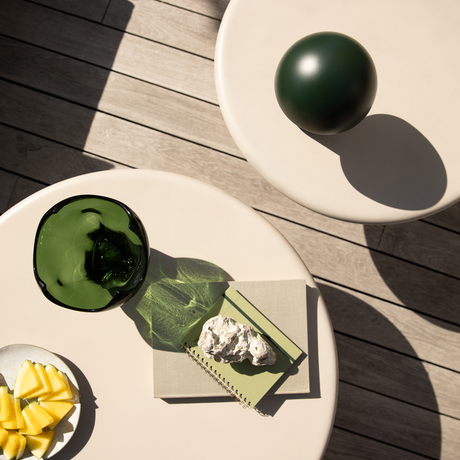 Överblick av två beige runda bord på en träterrass med en mörkgrön dekorativ kula, en grön glasbehållare som kastar skugga, två anteckningsböcker med en sten ovanpå och en liten tallrik med ananasklyftor i solljus.