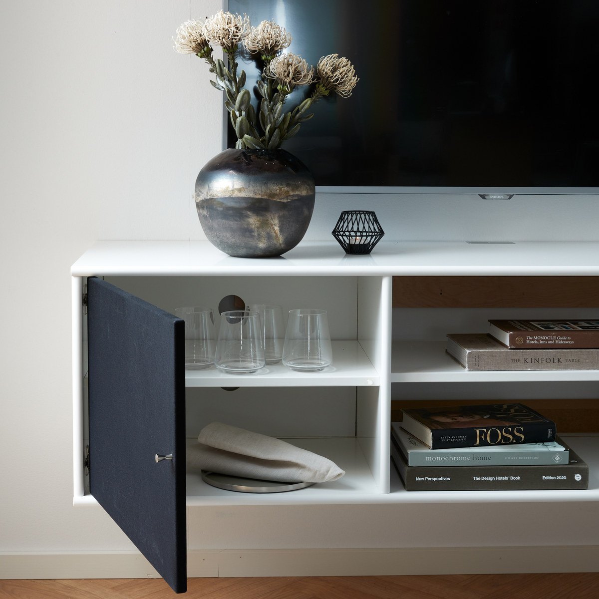 Vit mediabänk med TV, vas med torkade blommor och öppen lucka med glas; bredvid ligger en stapel designböcker: The Kinfolk Table, Monochrome Home, The Design Hotels Book, New Perspectives (Edition 2020) och Foss.