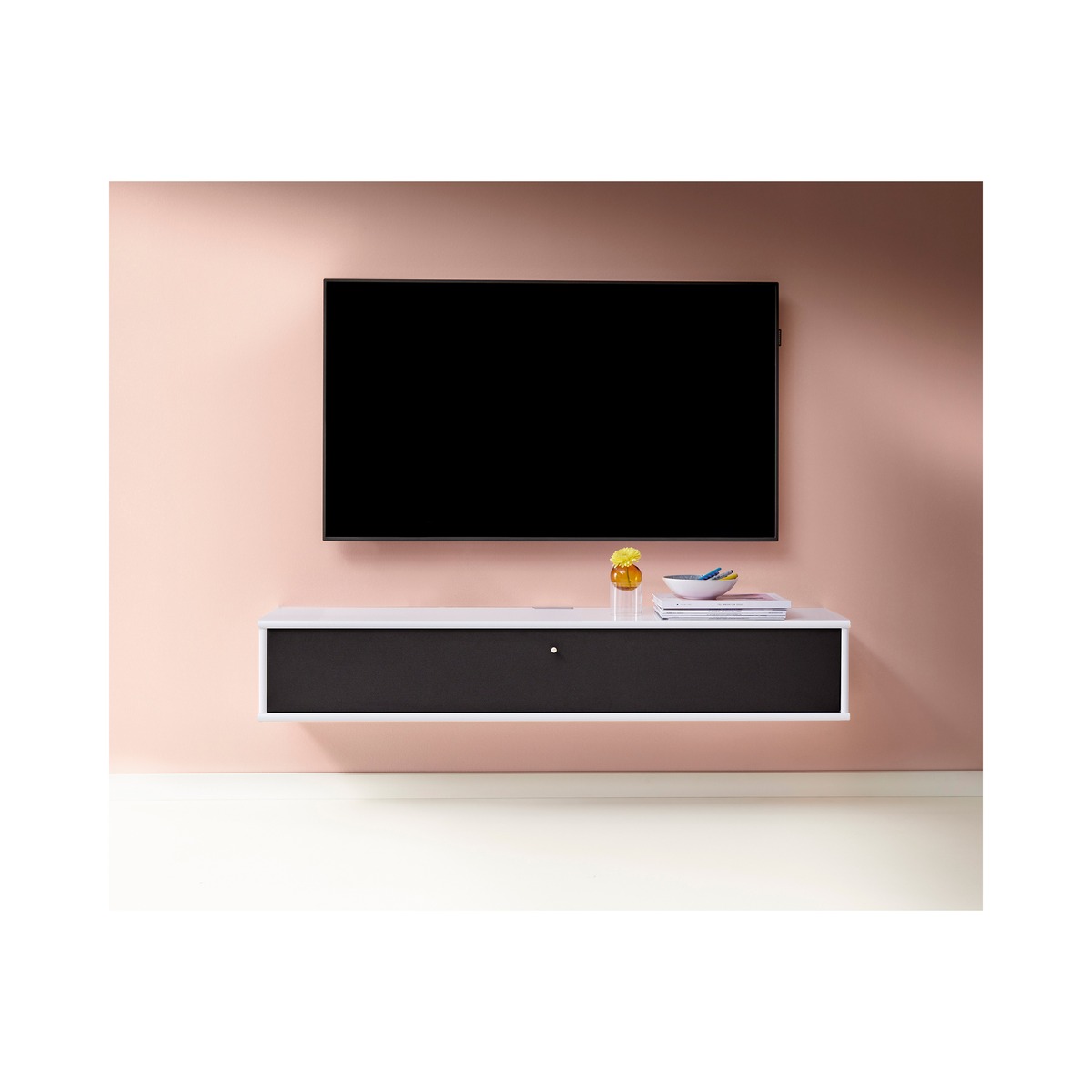 Väggmonterad TV ovanför svävande vit mediabänk med svart front; liten vas med gul blomma, skål och tidskrifter mot en mjukt rosa vägg.