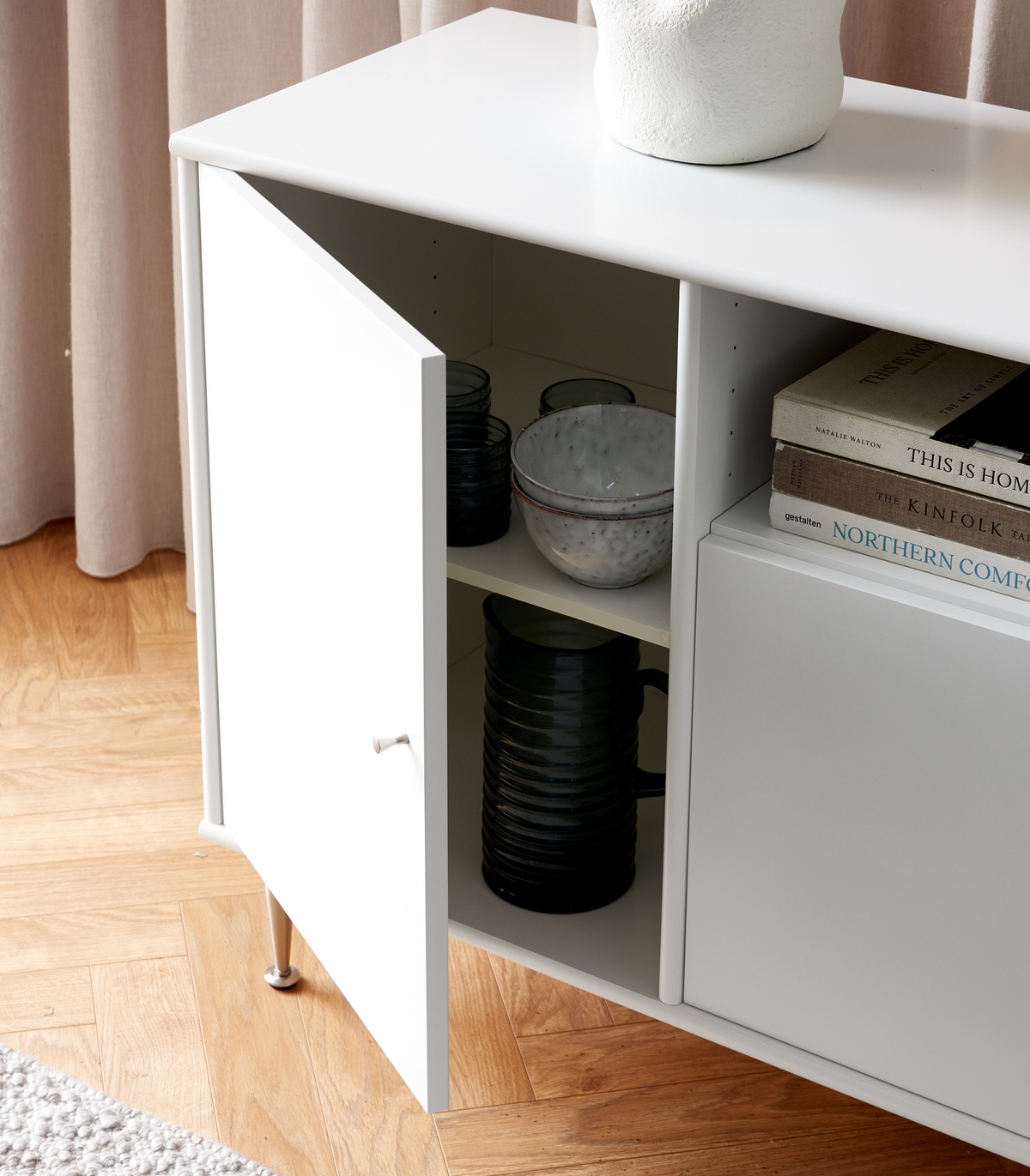 Vit sideboard med öppen dörr som visar glas och skålar; ovanpå står en vit vas och en stapel böcker med titlarna This Is Home, The Kinfolk Table och Northern Comfort.