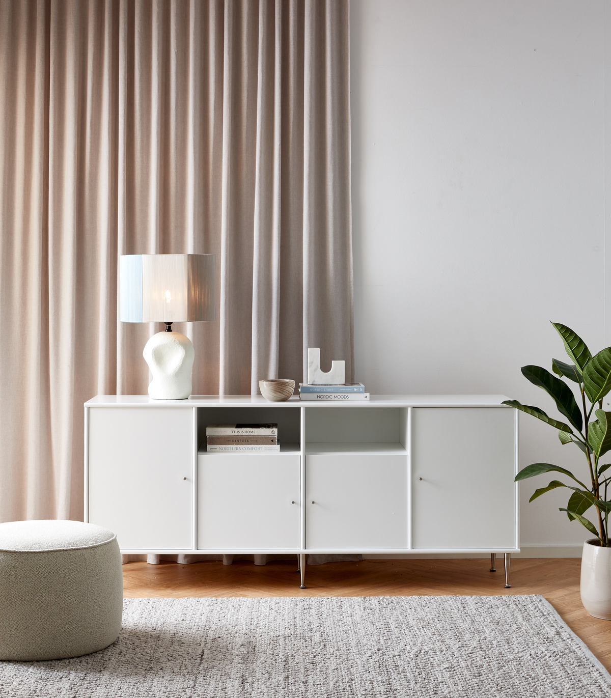 Ljust vardagsrum med vit sideboard, skulptural bordslampa, skål och böcker inklusive Nordic Moods, samt grön växt, beige gardiner och grå matta.