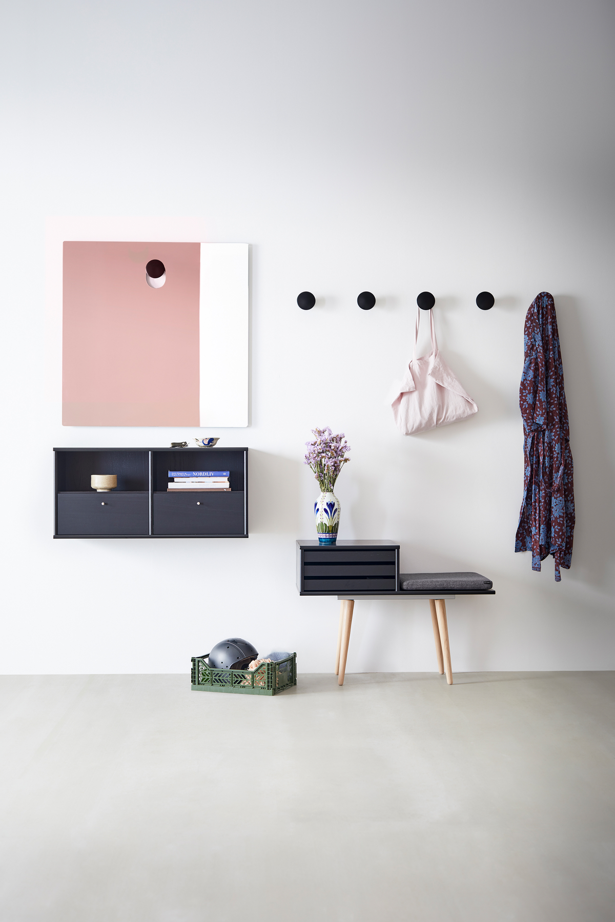Ljus entré/hall med svarta rundade väggknoppar, liten svart bänk med grå dyna, väggmonterade hyllor, rosa‑vit spegel och en vas med lila blommor samt grön korg på golvet.