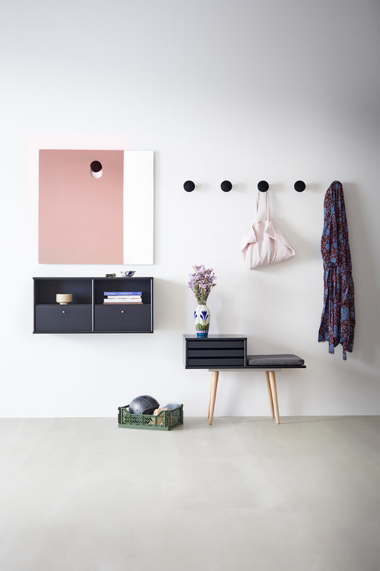 Ljus entré/hall med svarta rundade väggknoppar, liten svart bänk med grå dyna, väggmonterade hyllor, rosa‑vit spegel och en vas med lila blommor samt grön korg på golvet.