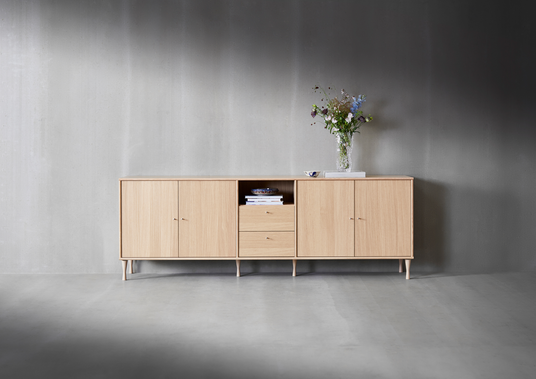 Sideboard i ljus ek med öppen hylla och två lådor, dekorerad med glasvas med blommor, liten skål och böcker mot en grå vägg.