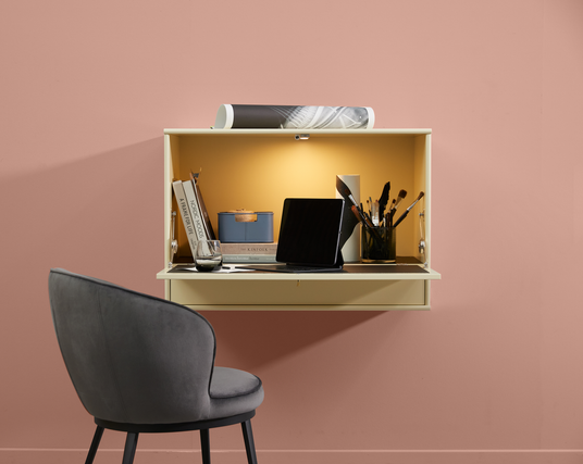 Vägghängd liten arbetsplats med surfplatta, böckerna A Frame for Life, Nordic Moods och The Kinfolk Home, penslar i burk och blå låda, varm belysning och grå stol mot rosa vägg.