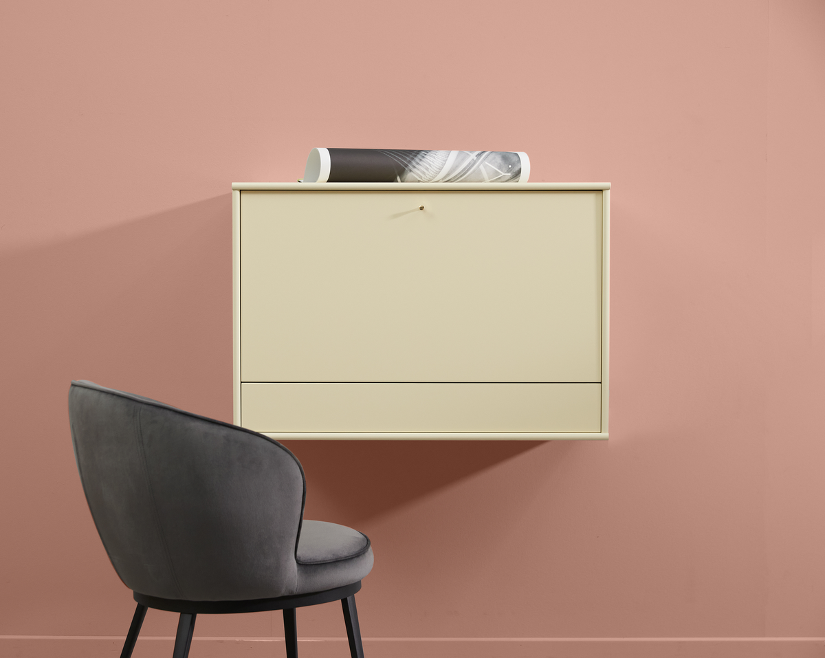 Beige vägghängt skåp mot rosa vägg med grå stoppad stol bredvid och en hoprullad affisch ovanpå.