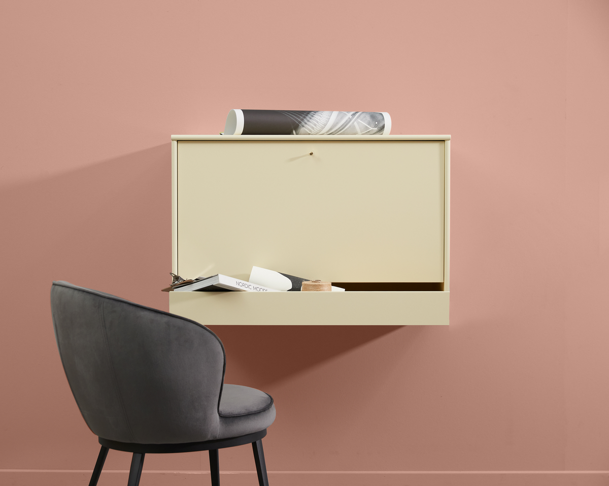 Väggmonterat beige skrivbord med öppen lucka, grå stol och en bok med titeln ”NORDIC MOOD”, mot en rosa vägg.