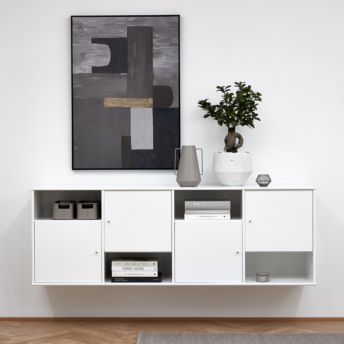 Vit vägghängd skänk med böcker (”Dior”, ”Live Beautiful”, ”Nordic Moods”, ”Kinfolk”), grå kanna och grön växt i vit kruka; ovanför hänger en abstrakt tavla i grå toner.