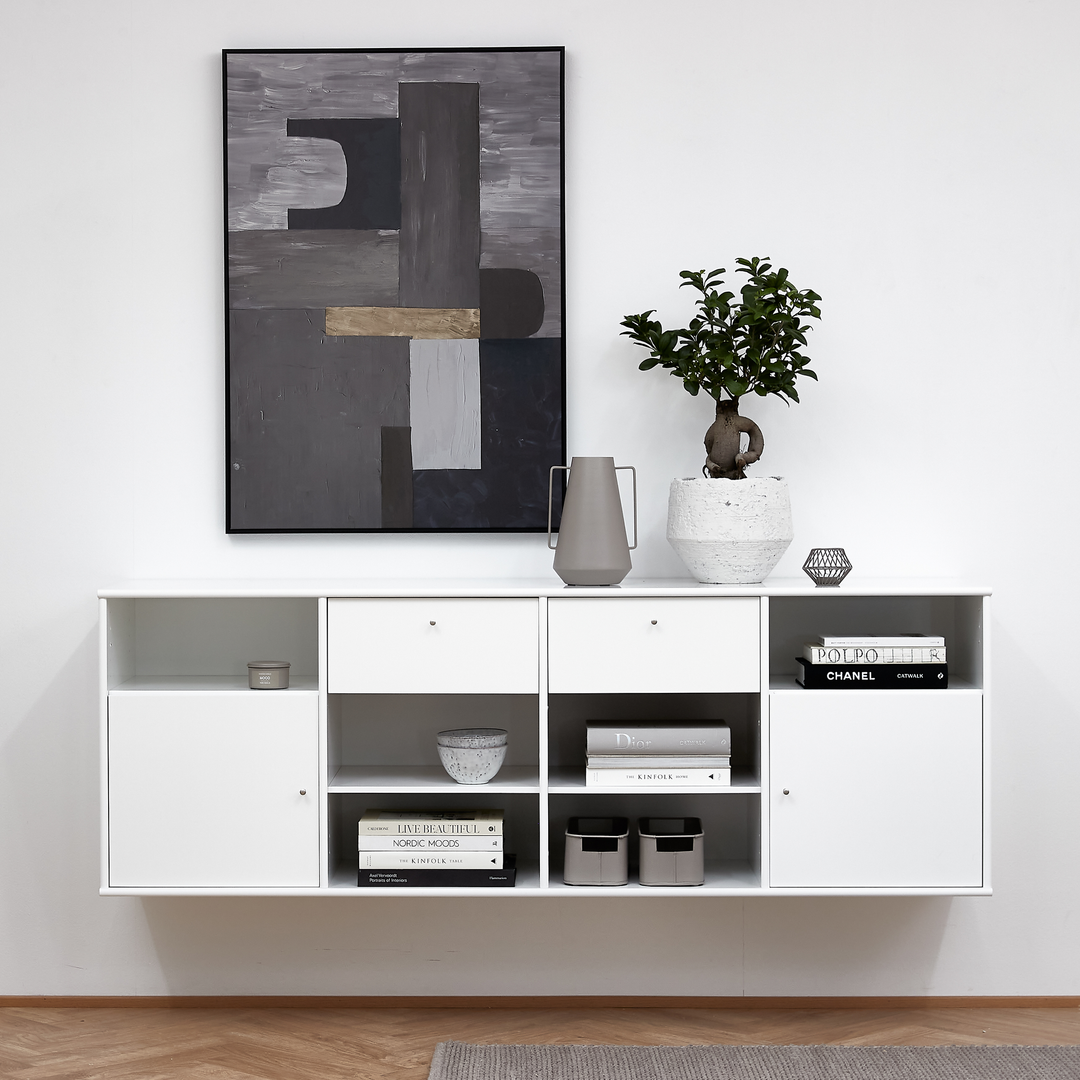 Minimalist vit vägghängd skänk med öppna hyllor med böcker – Chanel, Kinfolk, Live Beautiful, Polpo, Dior – samt skål och förvaringslådor; ovanför en abstrakt grå tavla och på toppen en grön växt i vit kruka och en grå kanna.