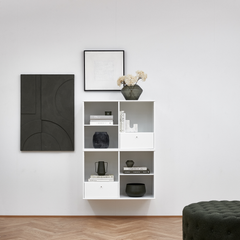 Väggmonterad vit hylla med böcker och keramik; titlarna ”Dior” och ”A FRAME APOLLO” syns; grön glasvas med torkade blommor ovanpå; två grafiska tavlor på väggen och en mörkgrön ottoman i förgrunden.