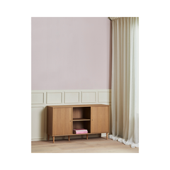 Låg sideboard i ek med öppna hyllor, mot ljusrosa vägg med panel och vita, skira gardiner.