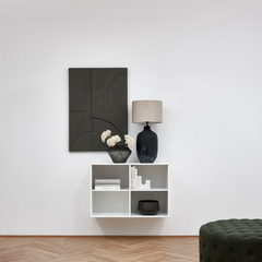 Minimalist vardagsrum med väggmonterad vit hylla, bordslampa med beige skärm, mörk vas med vita blommor och svart geometrisk väggkonst.