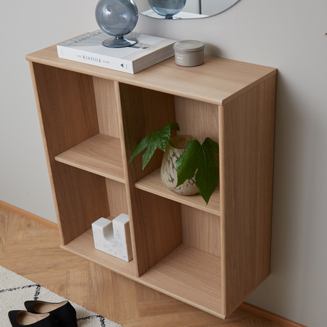 Väggmonterad hylla i ljus ek med boken The Kinfolk Home, en grå glasdekoration och en liten ljusburk ovanpå samt en grön växt i kruka och ett vitt marmorobjekt i facken.