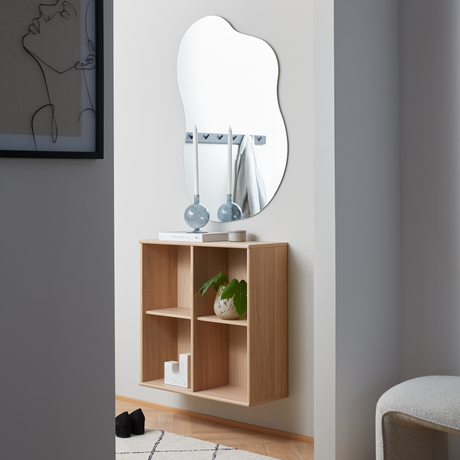 Minimalistisk hall med väggmonterad hylla i ek under en organiskt formad spegel, dekorerad med blå ljusstakar, en bok och en liten växt.