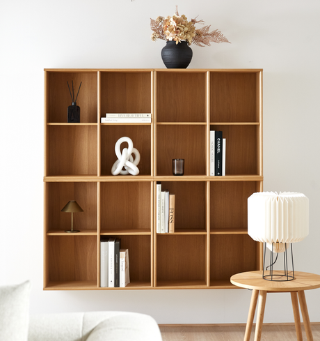 Vägghängd hylla i trä med böcker, CHANEL‑bok, skulptural vit knut, svart vas med torkade blommor samt liten bordslampa och en plisserad bordslampa på sidobord.