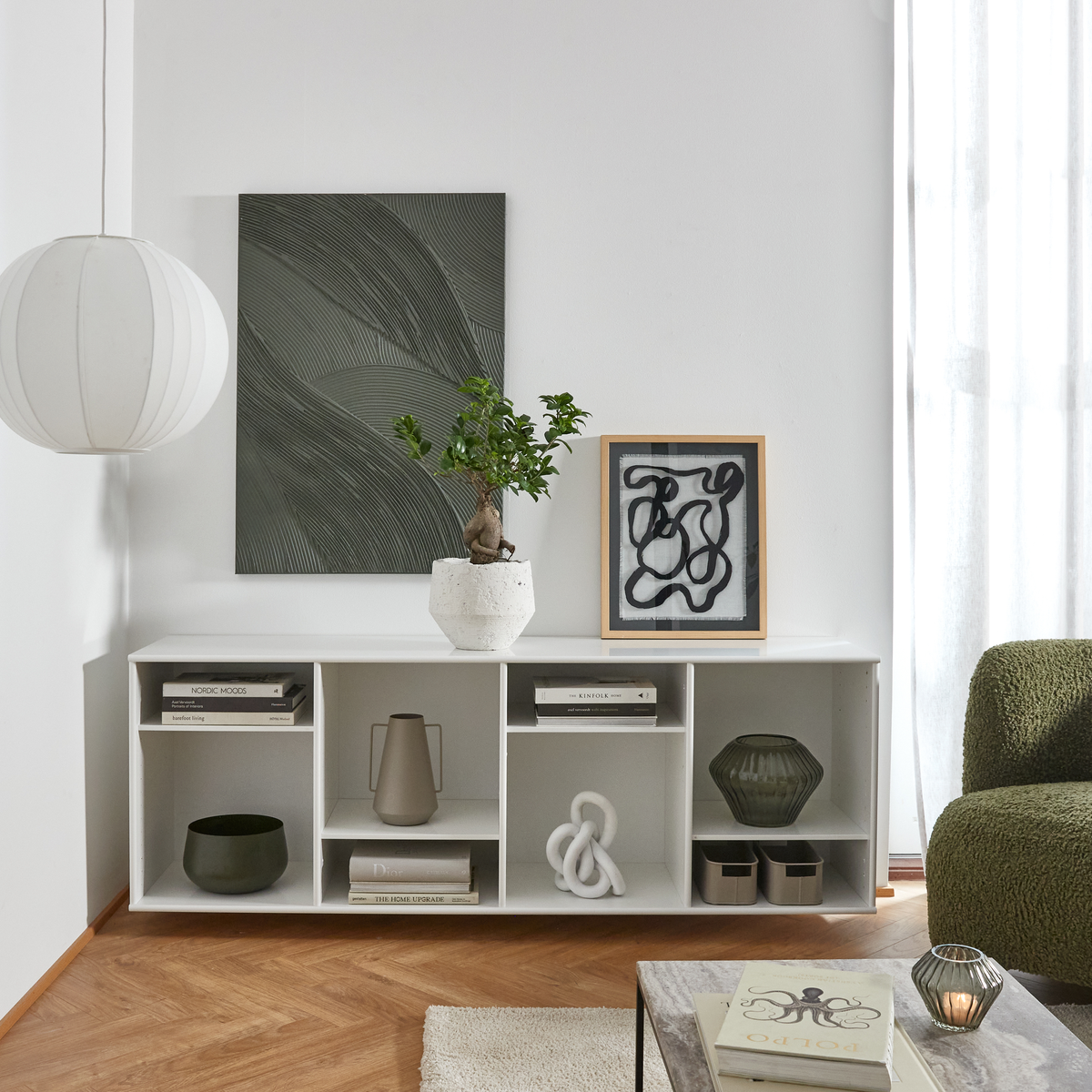 Minimalistiskt vardagsrum med vit förvaringsbänk, abstrakt väggkonst, bonsai i kruka och rund pendellampa bredvid grön soffa i dagsljus.