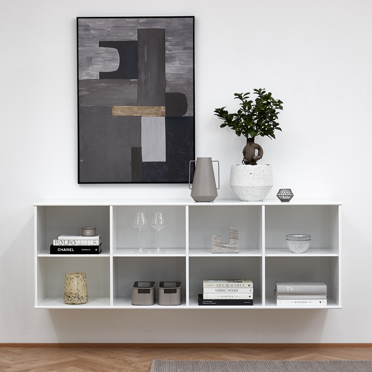 Vitt vägghängt hyllsystem med dekor; växt i kruka, grå karaff, glas, ljuslykta och abstrakt konst på väggen. Böcker inklusive Chanel, Nordic Moods, Live Beautifully och The Kinfolk.