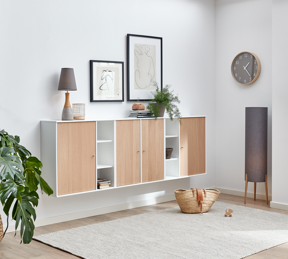 Skandinaviskt vardagsrum med vägghängda skåp i vitt och ljus ek, tavlor ovanför, växter, bordslampa, golvlampa, väggklocka och flätad korg på en ljus matta.
