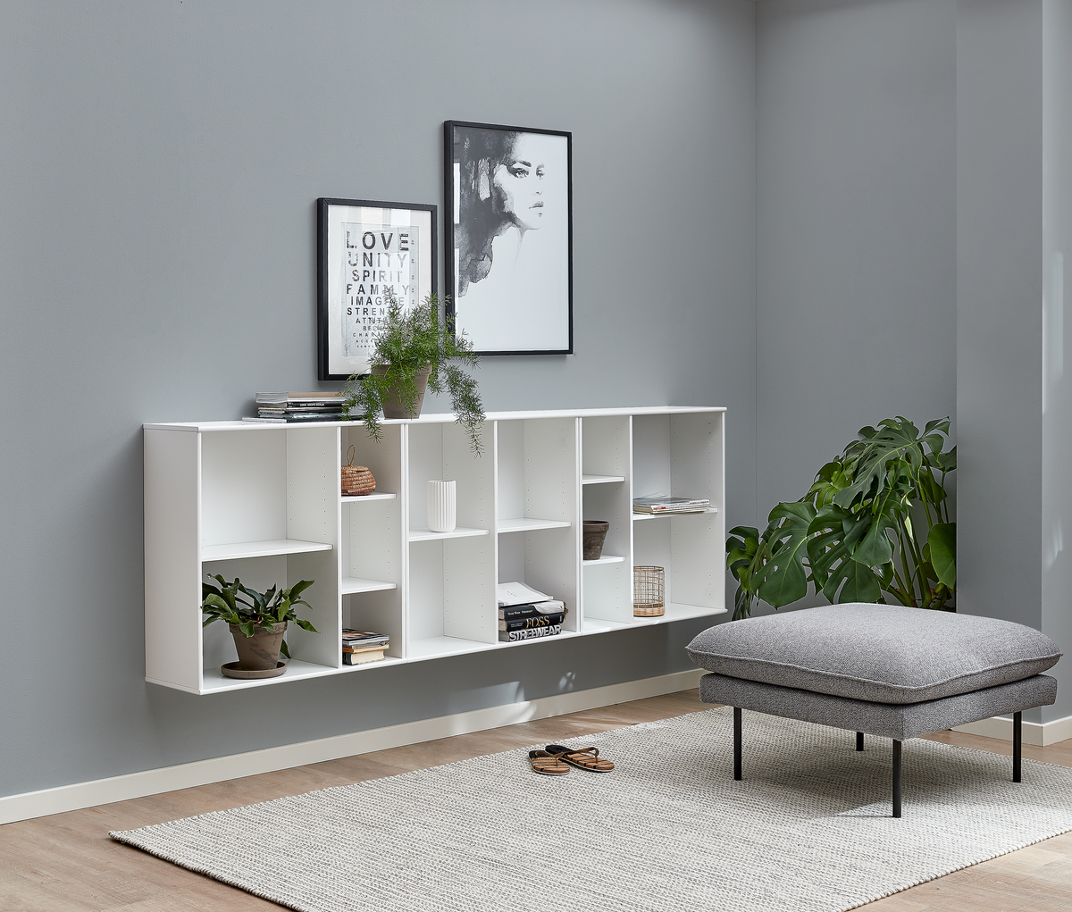 Minimalistiskt vardagsrum med väggmonterad vit hylla, två svartvita affischer med “LOVE UNITY SPIRIT FAMILY IMAGE STRENGTH”, gröna växter och en grå fotpall på ljus matta.