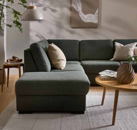 Mörkgrön hörnsoffa med beige kuddar i vardagsrum; träbord med skulpterad vas och böckerna ”Billedkunstens” i mjukt dagsljus, skandinavisk stil.