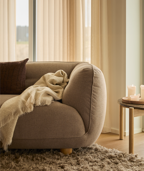 Närbild av en varm, neutral hörna i vardagsrummet: brun soffa med krämfärgad pläd och kudde, lång luggmatta och litet träbord med tända ljus framför beige gardiner i dagsljus.