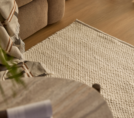 Närbild av beige, texturerad matta bredvid soffa med rutigt pläd på trägolv i ett mysigt vardagsrum.