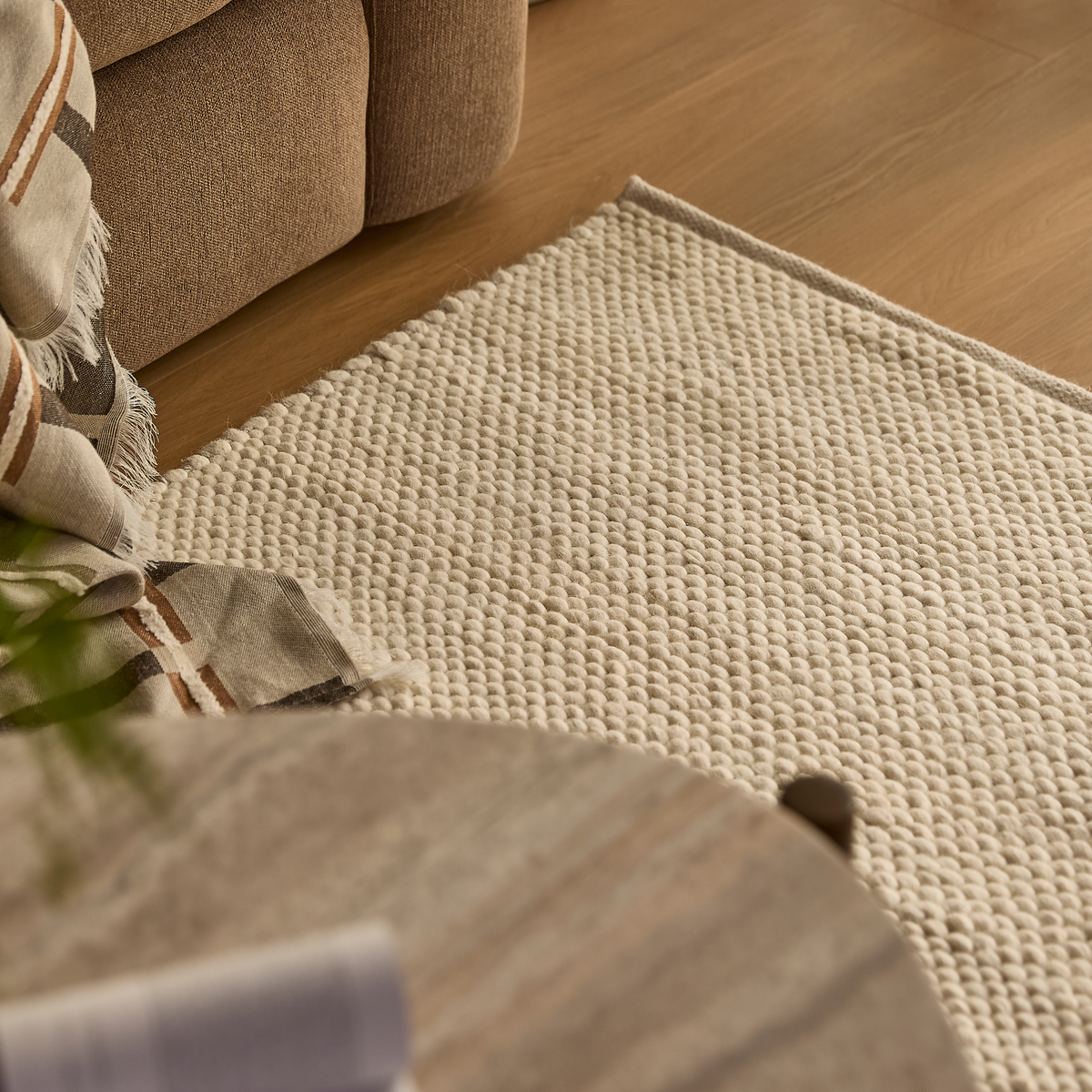 Närbild av ljus, texturerad matta bredvid beige soffa, med randig pläd och trägolv i varmt dagsljus.