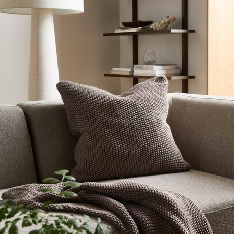 Beige soffa med grovstickad brun kudde och matchande pläd; hylla, lampa och grön växt i ett ljust vardagsrum.