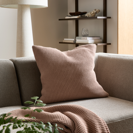 Beigefärgad soffa med en chunky-stickad puderrosa kudde och matchande pläd; golvlampa och hyllor med böcker och dekoration i ett lugnt vardagsrum.